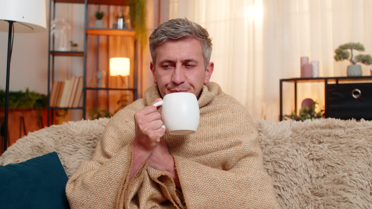 Young man wrapped in blanket with tea looking sick recovering from cold with mild seasonal symptoms