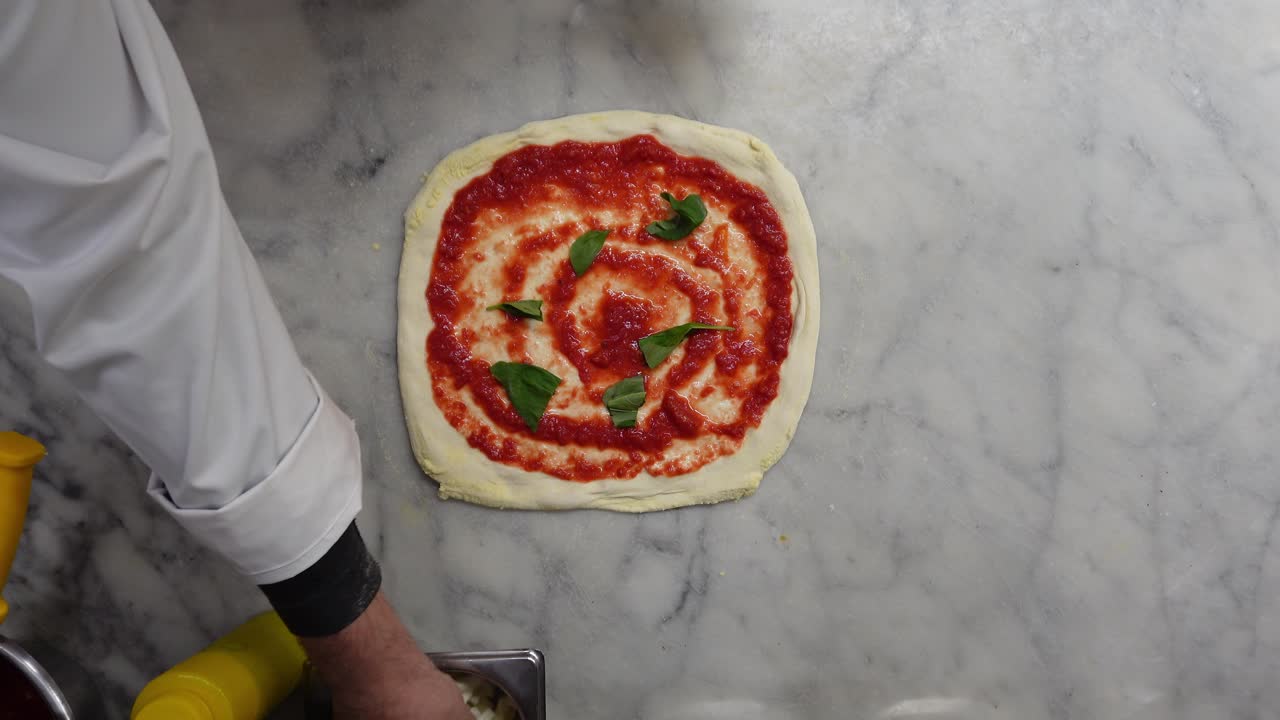 Vista de cima de um chef italiano colocando molho de tomate, manjericão e mussarela na massa de pizza para fazer uma pizza Margherita.