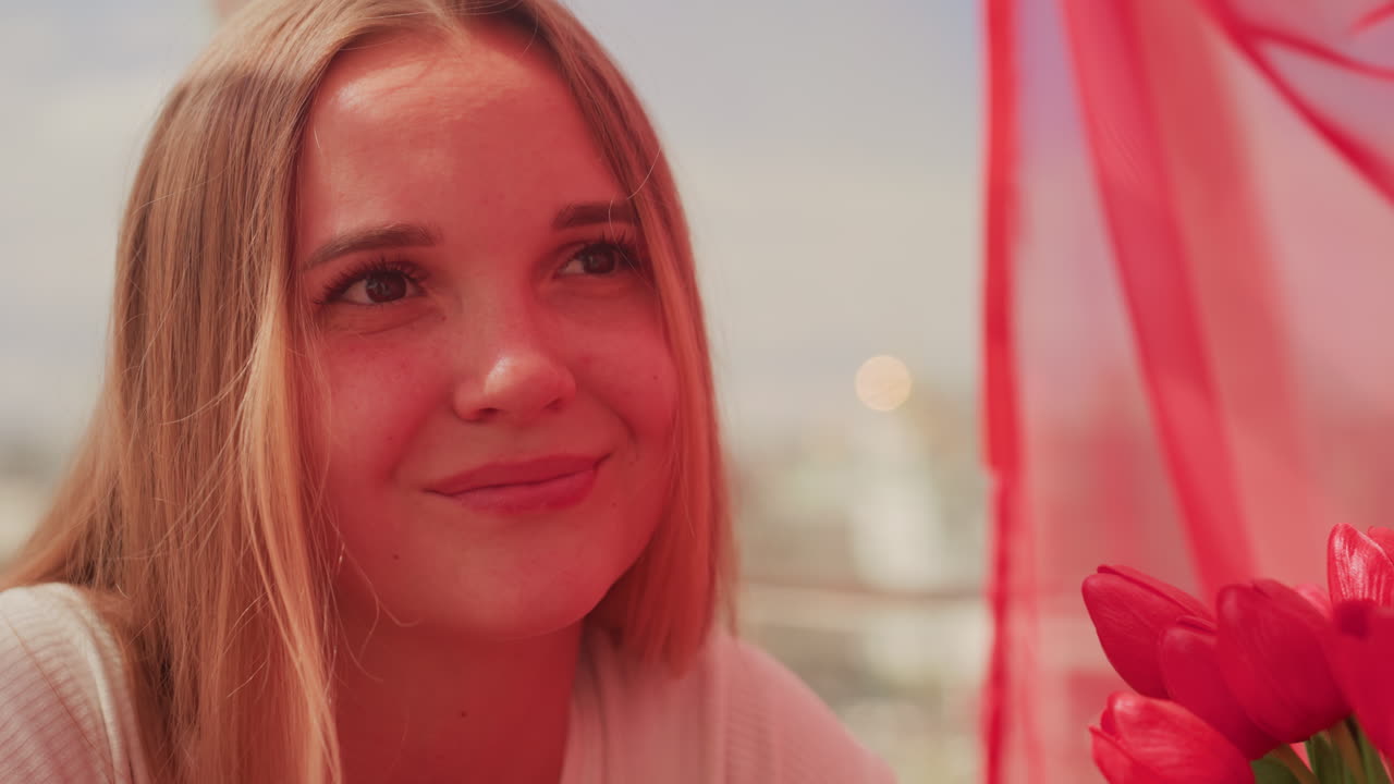 Ung, hvid kvinde smiler på en balkon, hendes partners hånd rækker hende en tulipan, rødt, transparent gardin, gyldent sollys, byens skyline er sløret, intimt og romantisk udtryk, blød bokeh, ærligt nærbillede