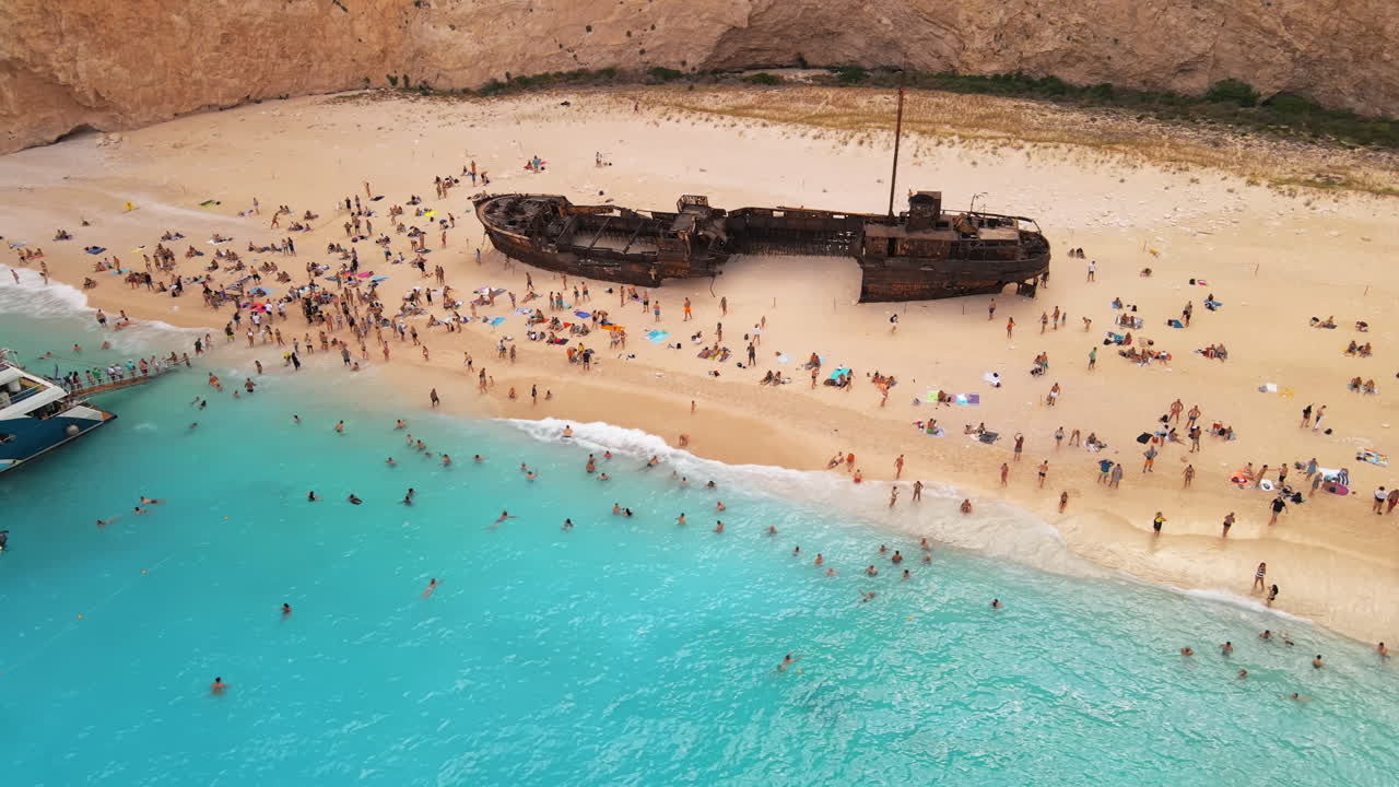 Aerial drone view of the Navagio beach on the Ionian Sea coast of Zakynthos, Greece. Moored boats and resting people, rusty ship, blue water