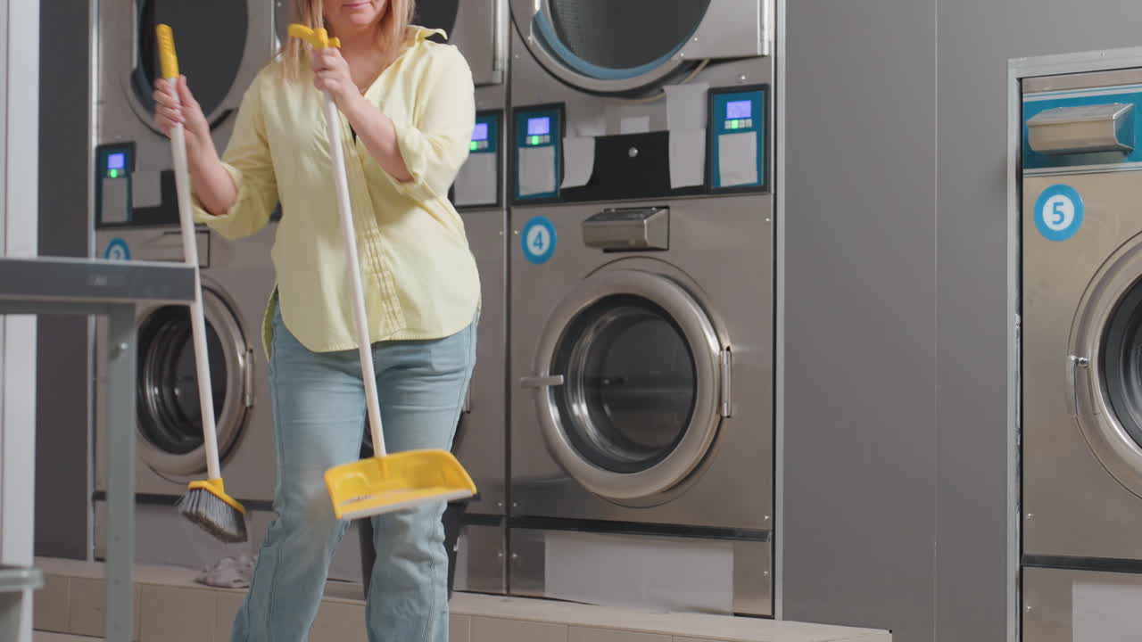 Joyful business woman dances while sweeping near industrial washer in laundromat, holding broom stick and dust pan, cleaning lint spilled from filter drawer on floor during maintenance routine