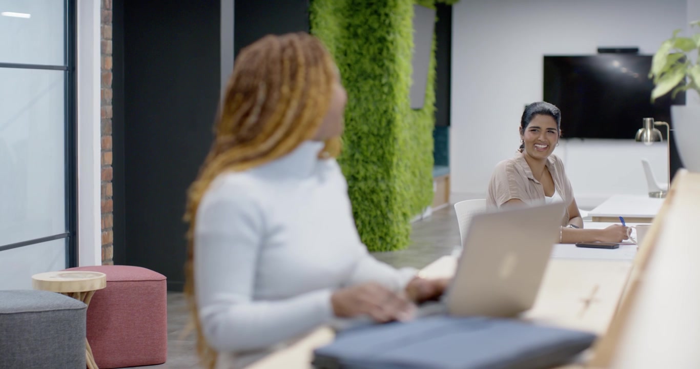 Diverse female coworkers, Indian typing on laptop at counter, turning after coworker smiling hello