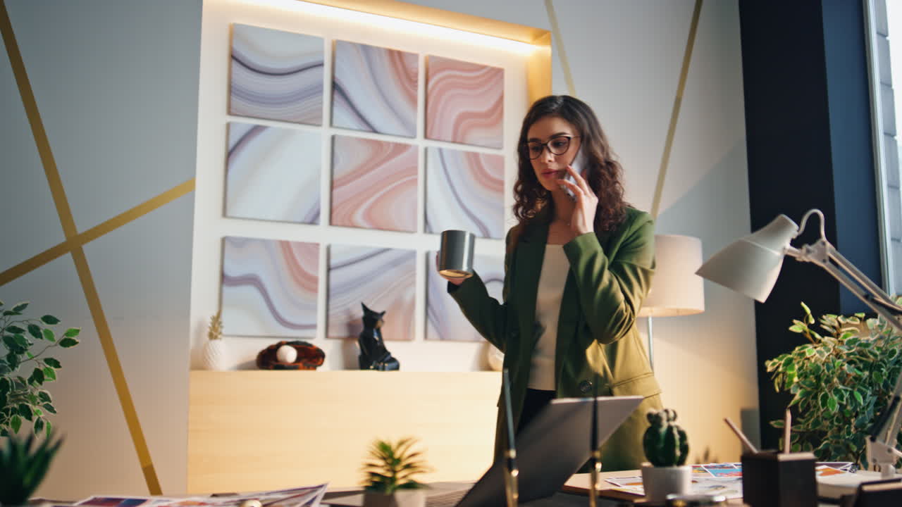 여자 상사가 커피 컵을 들고 사무실에서 전화를 하고 있다. 매니저가 술을 마시며 전화하고 있다.