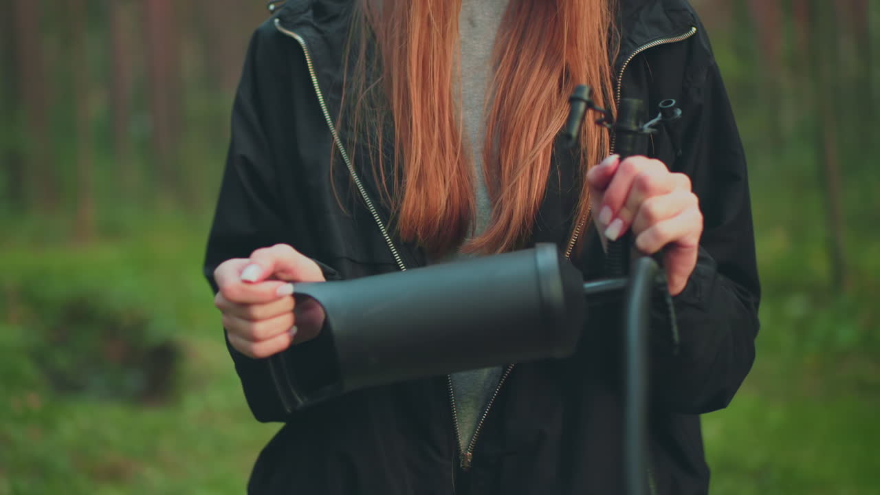 Close up of woman with long reddish hair wearing black jacket and grey shirt while holding and pumping nozzle with both hands, zipper partially open, in green forest environment