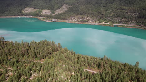 Atemberaubende-Rotierende-Luftaufnahme-Eines-Grünen,-Blaugrünen,-Türkisfarbenen-Sees-Mit-Einer-Wunderschönen-Küste-In-Norwegen,-Spiegelungen-Auf-Dem-Ruhigen-Wasser,-Norwegische-Forststraße,-Etwas-Verkehr-Im-Hintergrund,-Skandinavien