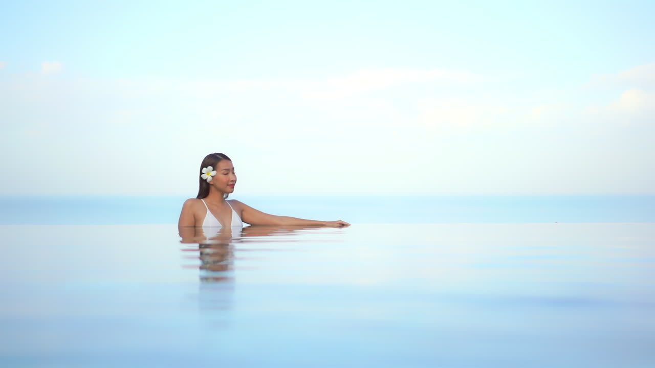Junge, schöne Frau im Badeanzug lehnt am Rand eines Infinity-Pools mit verschwommenem Ozean und blauem Himmel im Hintergrund.