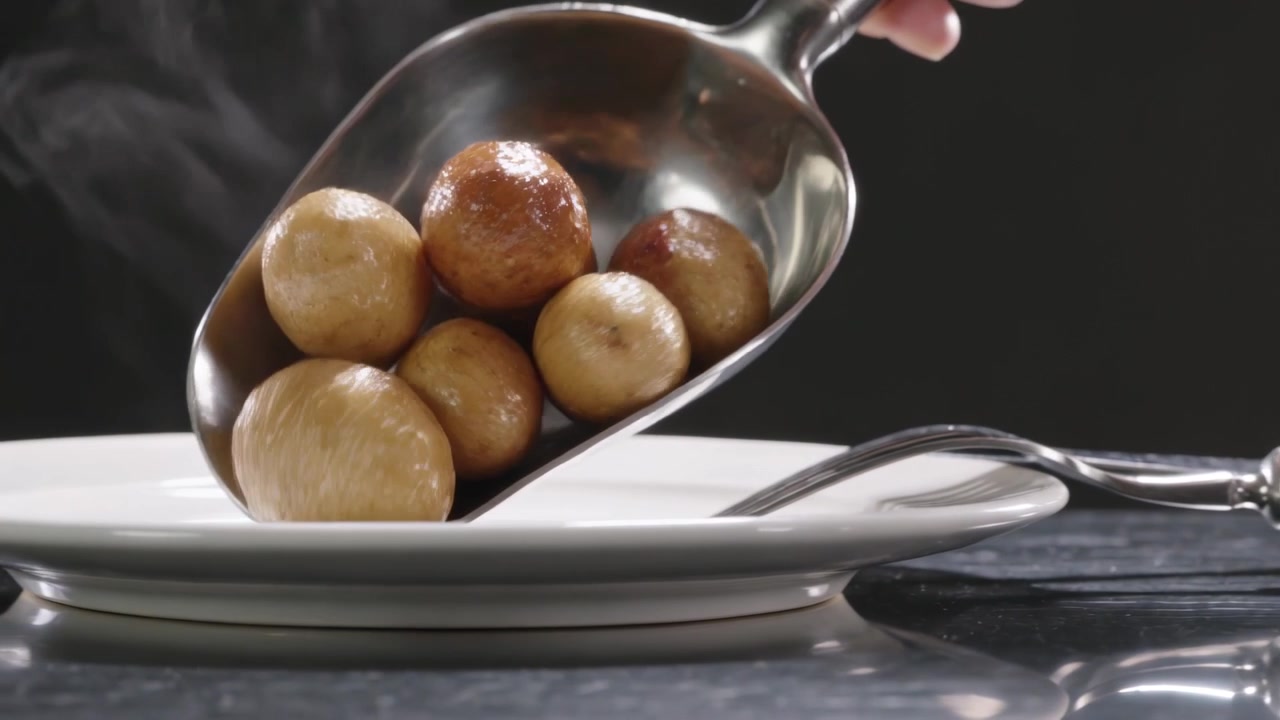 Entering metal scoop, hand tilting, plating steaming potatoes on white plate at counter, fork right