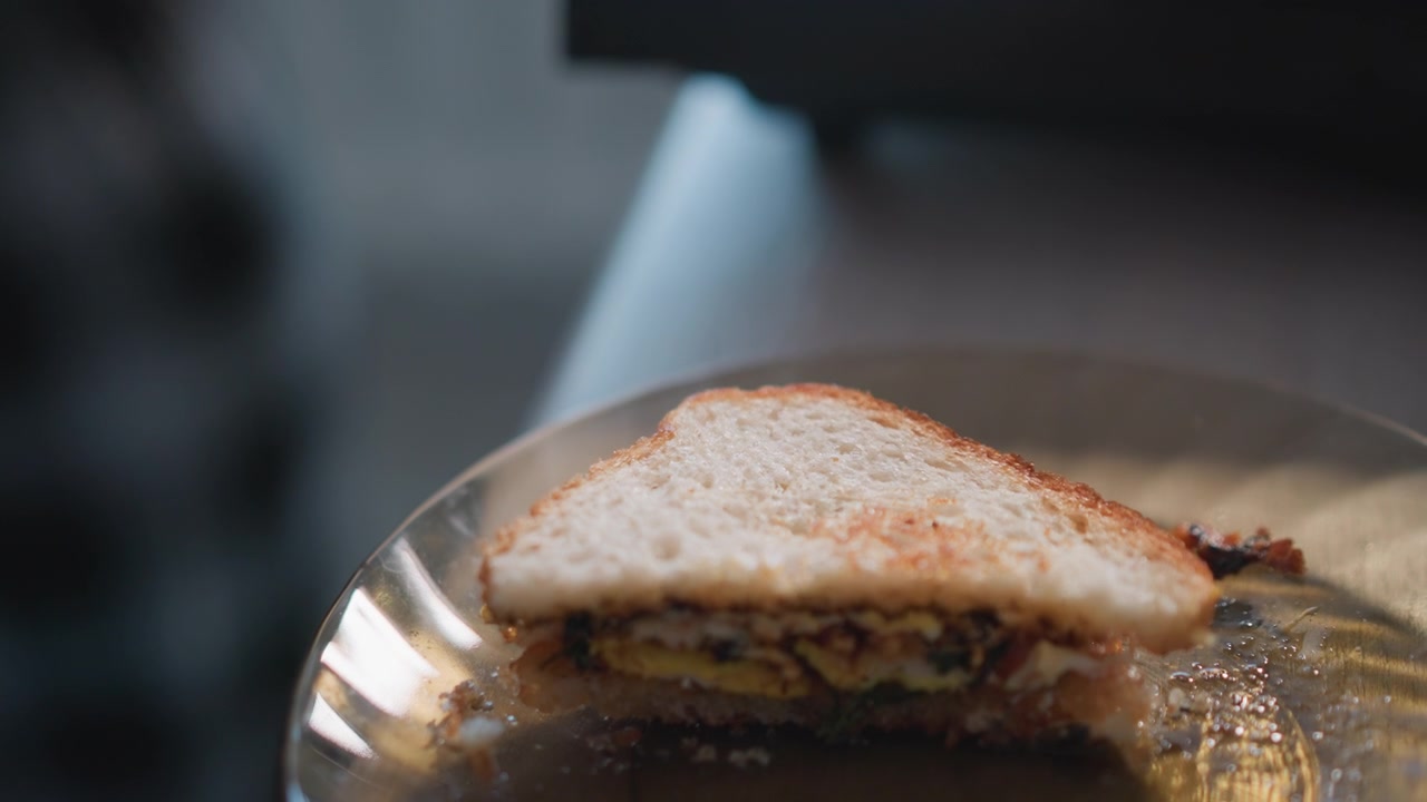 Primo piano di sandwich tostato su piatto, crosta dorata e lucentezza del burro, piatto di vetro riflettente, sfondo sfocato della cucina, profondità di campo ridotta, atmosfera tranquilla di tarda notte, briciole e texture rustica