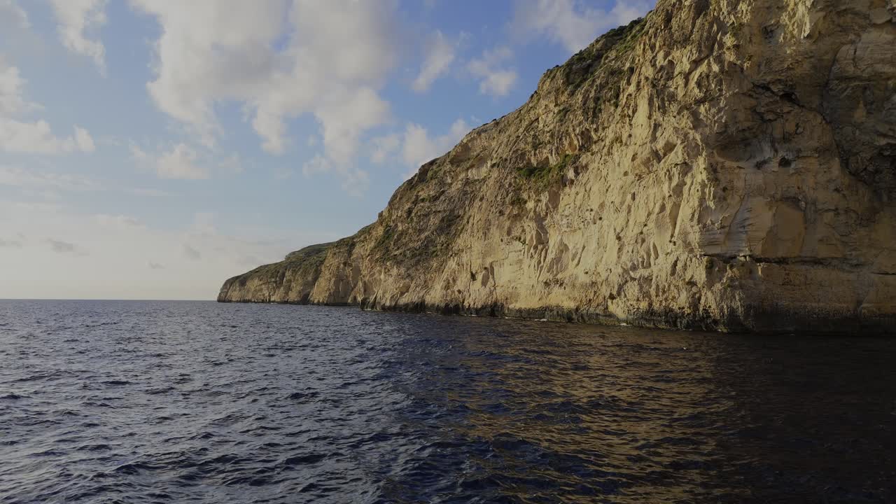 Un volo dinamico di un drone lungo le scogliere di Xaqqa, illuminate dalla calda luce del tramonto, con il mare calmo sottostante e un cielo luminoso con soffici nuvole bianche