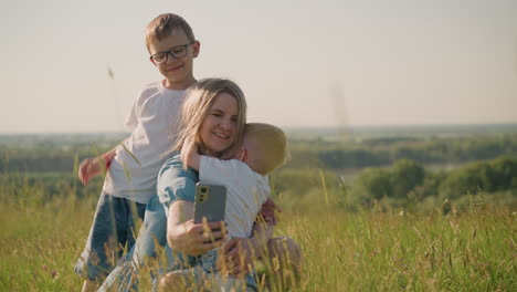 a woman in blue dress sits on the grass, smiling as takes a picture with her smartphone. her two sons, are with the older son is standing beside while the younger one hugs her closely