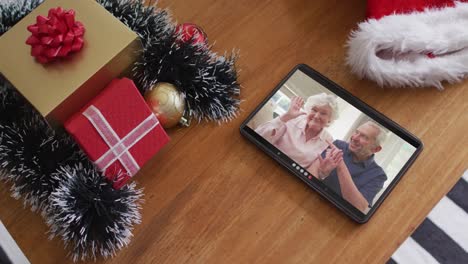 una pareja caucásica mayor sonriente saludando en una videollamada de navidad en una tableta