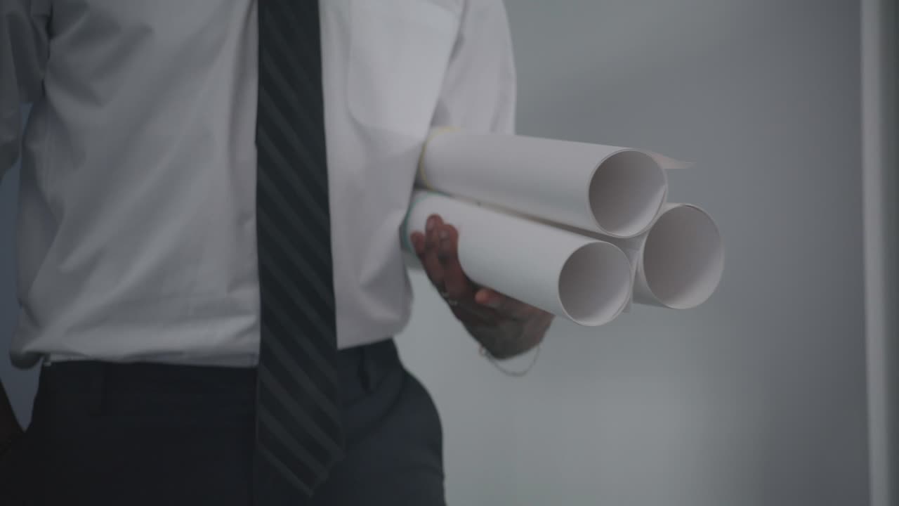 A man dressed in business attire walks through an office while carrying rolled blueprints. A concept of architecture, engineering, project planning, and corporate professionalism