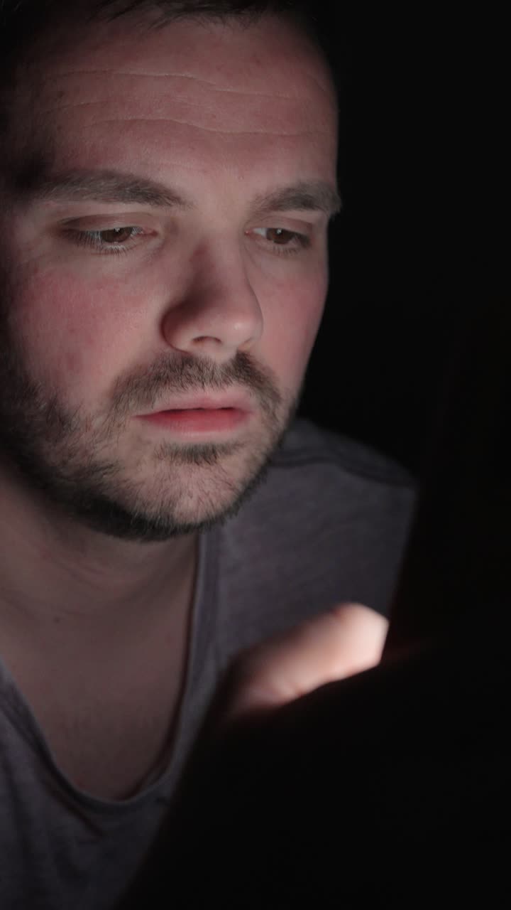 Primo piano verticale di un uomo che digita sul suo smartphone al buio, illuminato dalla luce dello schermo, che mostra messaggistica notturna, focus digitale e una scena di vita interna d'atmosfera.