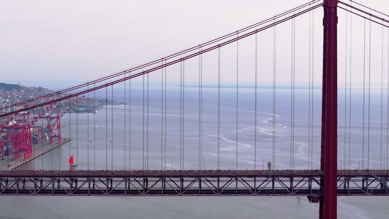 Side-on aerial drone shot of half marathon and 10K long-distance running event in Lisbon, Portugal, Europe. Runners crossing the iconic famous red 25th April suspension bridge