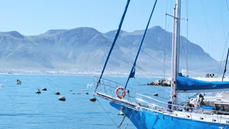 a blue sailing yacht, docked in harbor, on a calm day, with buoys and mountains in the distance