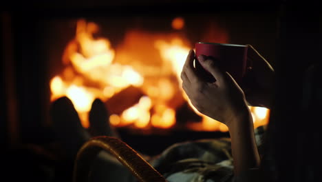 Tee-Am-Kamin---Weibliche-Hände-Mit-Einer-Roten-Tasse-Auf-Dem-Hintergrund-Des-Kamins,-Wo-Das-Feuer