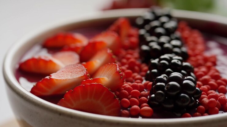 Primo piano angolato dal basso di una ciotola di ceramica riempita con guarnizioni di frutta disposte ordinatamente: fragole a fette sovrapposte in primo piano, un denso grappolo di piccole perle sferiche rosse nella zona centrale e elementi verticali.