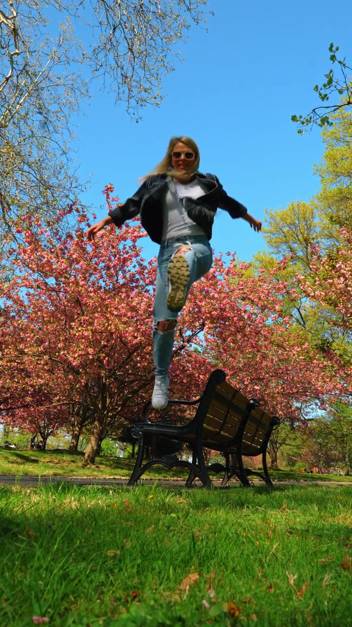 Movimento sfocato di una donna che salta in un parco di New York. Inquadratura dal basso con sfocatura di movimento di una donna che salta davanti a una panchina del parco e alberi di ciliegio in fiore rosa