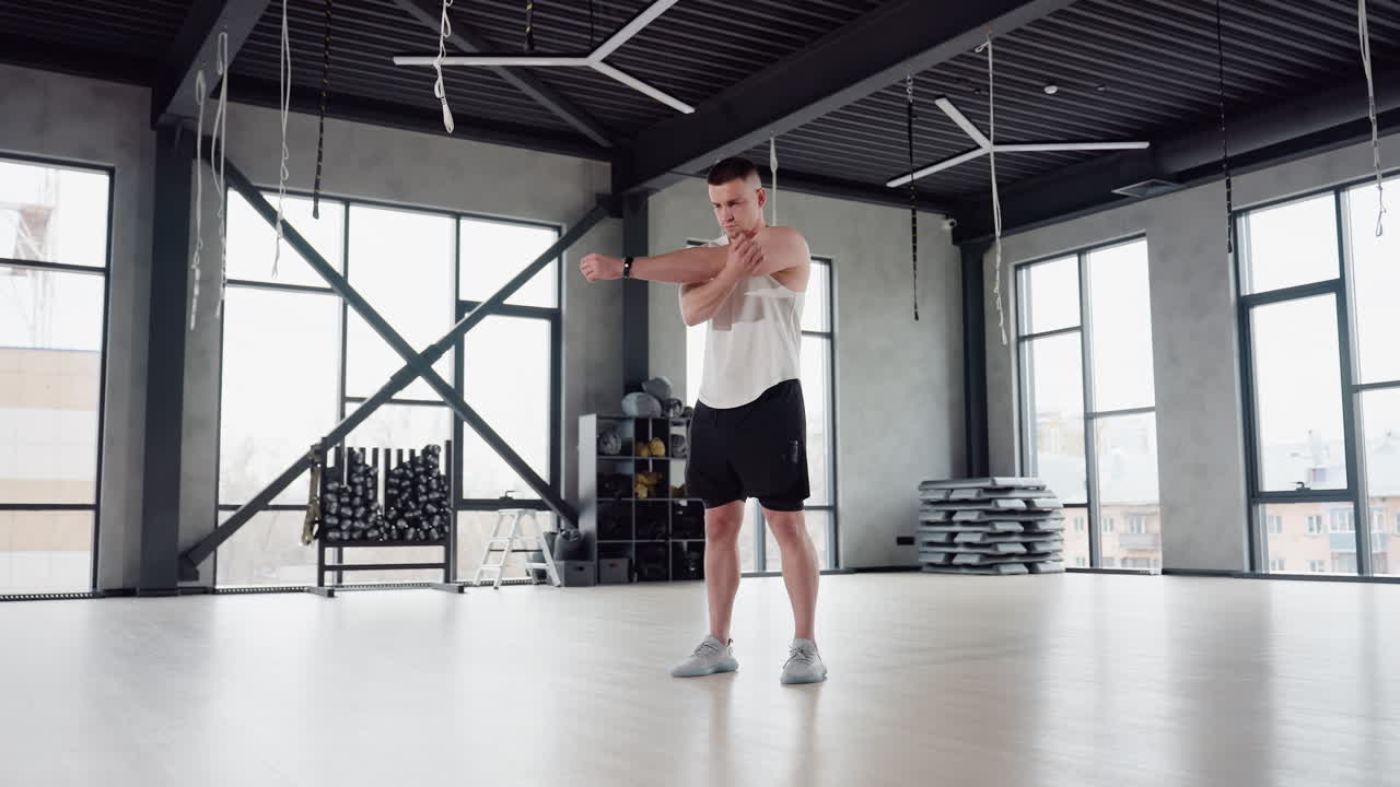 Fitness enthusiast performing warm up in spacious gym room with raised arms, preparing for dynamic workout, natural light through large windows, equipment arranged in background conveying energy
