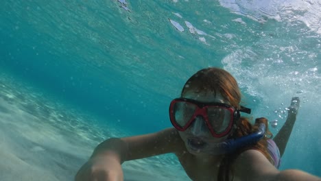 podwodne selfie małej rudowłosej dziewczynki z maską do nurkowania i fajką podnoszącą piasek ręką z dna morskiego i pływającą w czystej wodzie oceanu