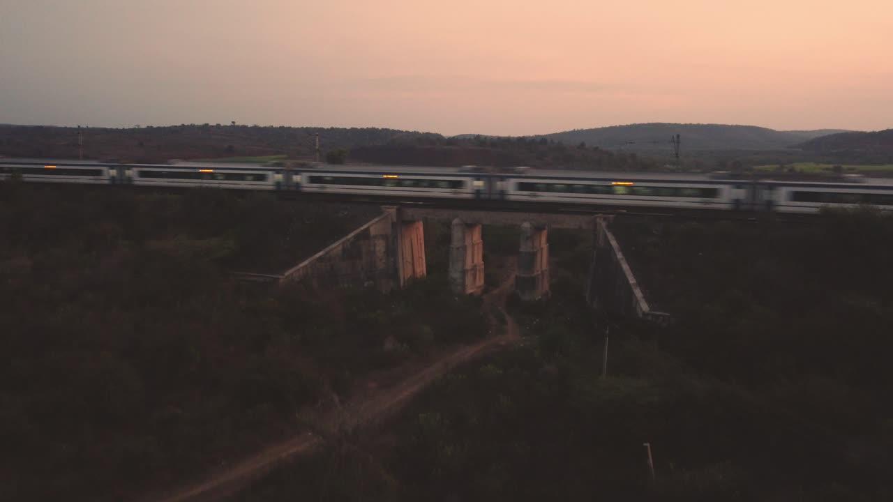 tomada aérea de un tren de vande bharat de los ferrocarriles indios que se mueve rápidamente en un viejo puente ferroviario de hormigón con densas colinas forestales en el fondo durante la tarde