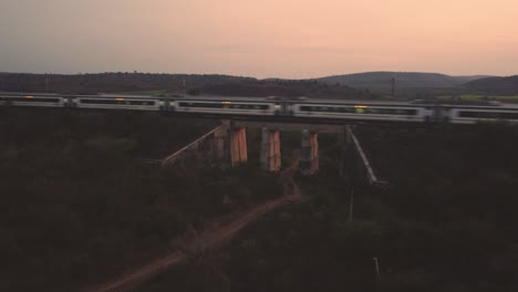 Aerial-drone-shot-of-an-Indian-railways-vande-bharat-train-moving-fast-on-an-old-concrete-Railway-bridge-with-dense-forest-hills-in-background-during-late-evening-time