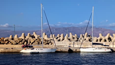 two sailboats docked near a concrete breakwater with mountains in the background on a sunny day