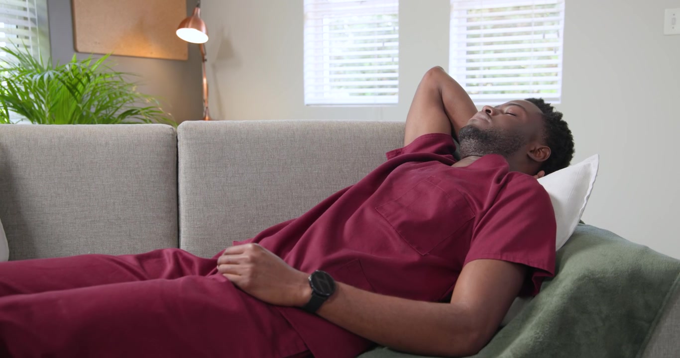 African American man in scrubs finishing shift, scrolling smartphone then resting on sofa at home