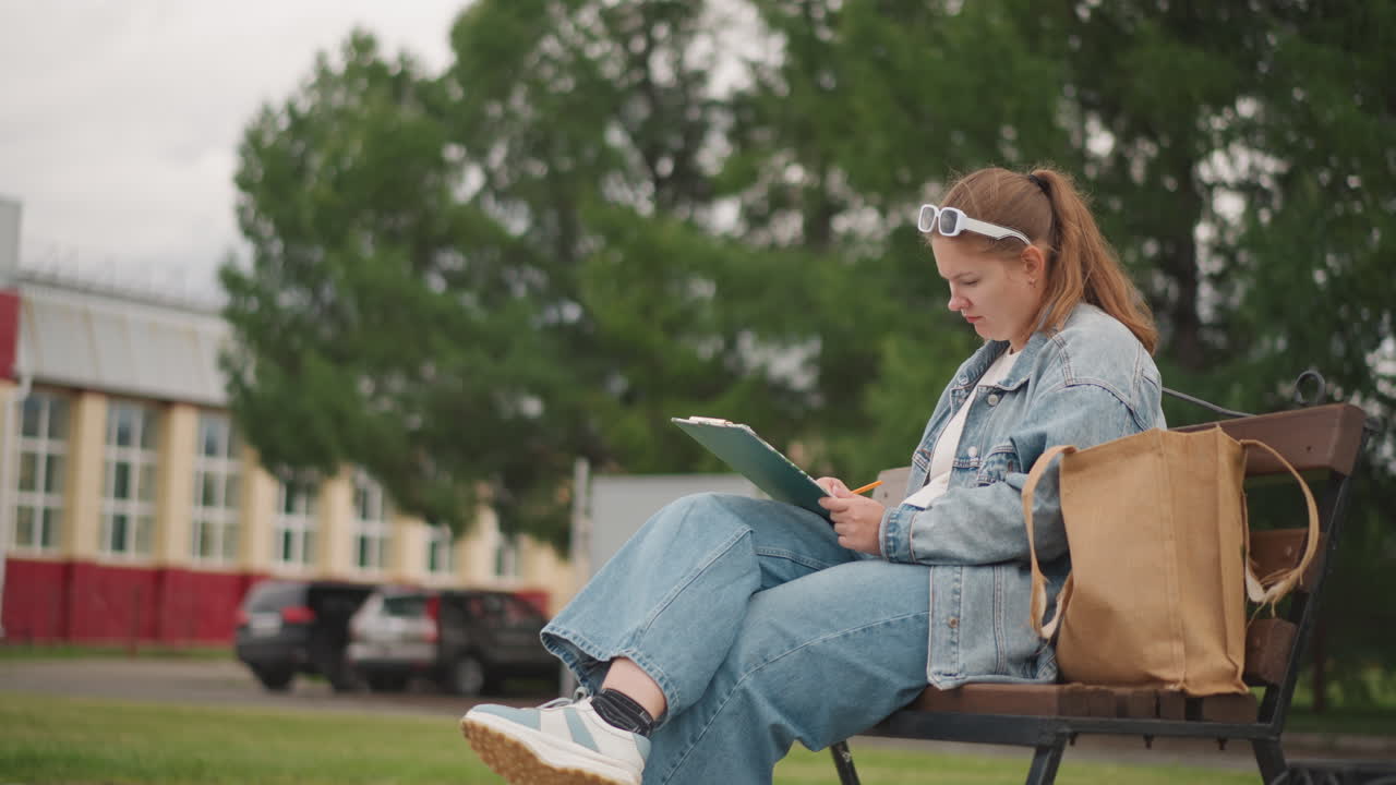 Donna bianca seduta su una panchina che scrive su un quaderno, abbigliamento in denim, occhiali da sole sulla testa, borsa tote accanto, strada del campus e alberi sullo sfondo, espressione concentrata, atmosfera di studio, primi piani di matita impugnata.