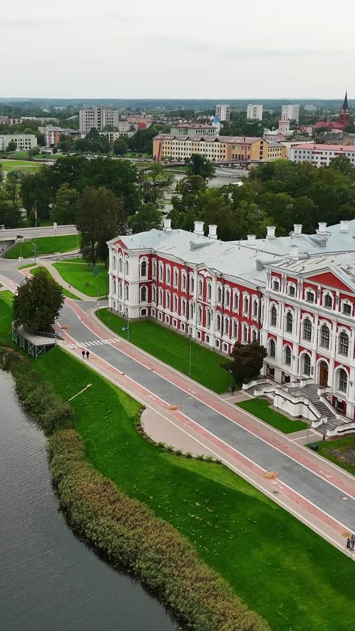 Vertical aerial perspective reveals Jelgava Palace's magnificent white and terracotta-red Baroque architecture, set majestically on a river island surrounded by manicured grounds and embankments
