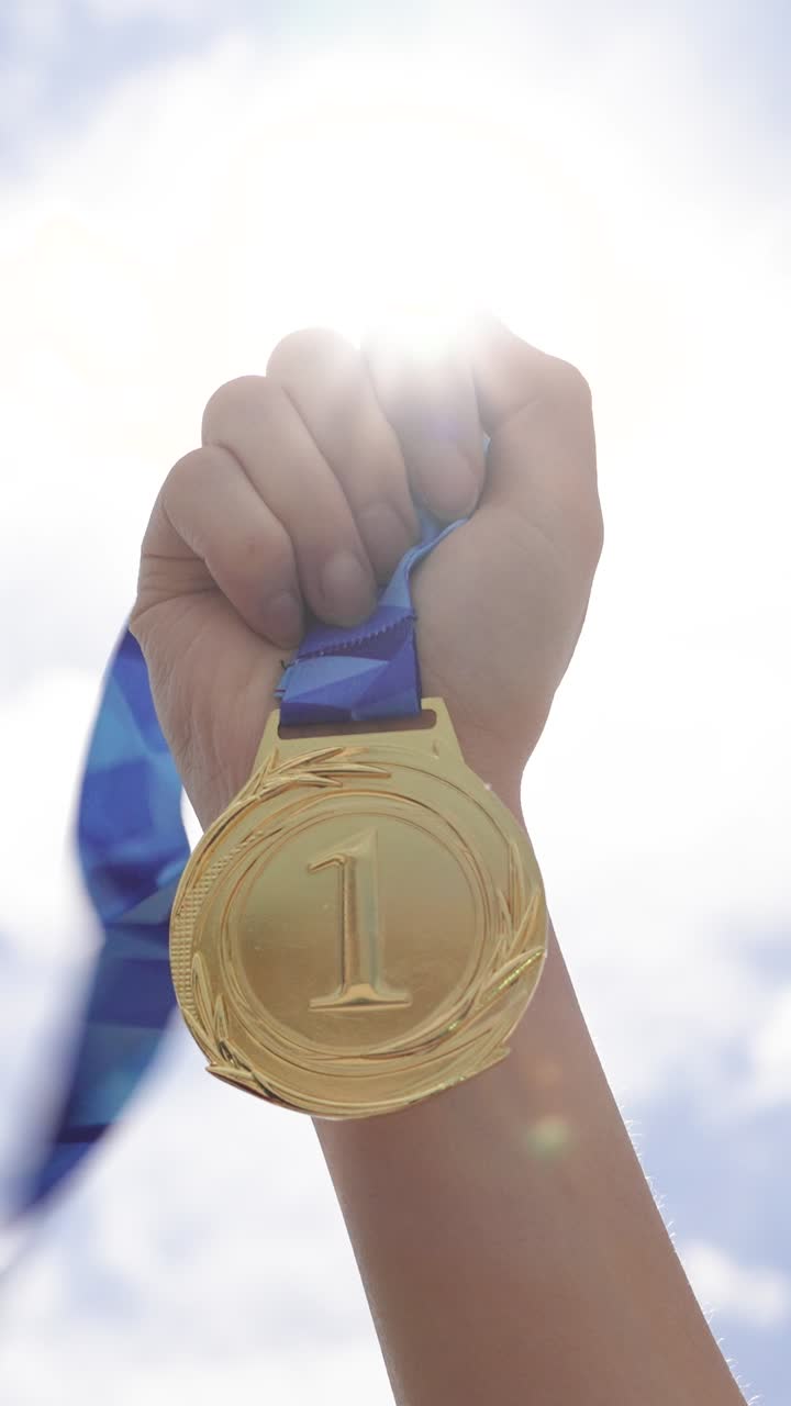 Mano che tiene una medaglia d'oro contro uno sfondo di cielo aperto in primo piano. Concetto di realizzazione, successo, vittoria, riconoscimento e motivazione alla vittoria.