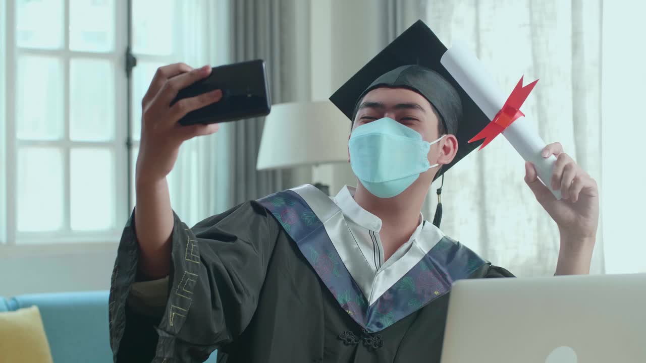Giovane uomo asiatico che indossa una mascherina protettiva, si scatta un selfie mentre indossa una toga e un tocco da laureato, con in mano un certificato universitario in salotto.