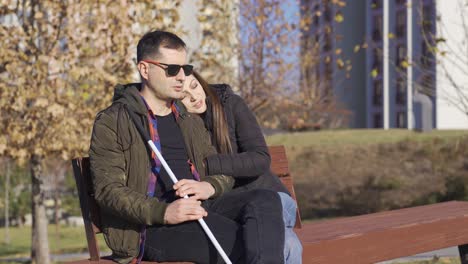 blind man and his wife sitting on a bench. his wife hugs him.