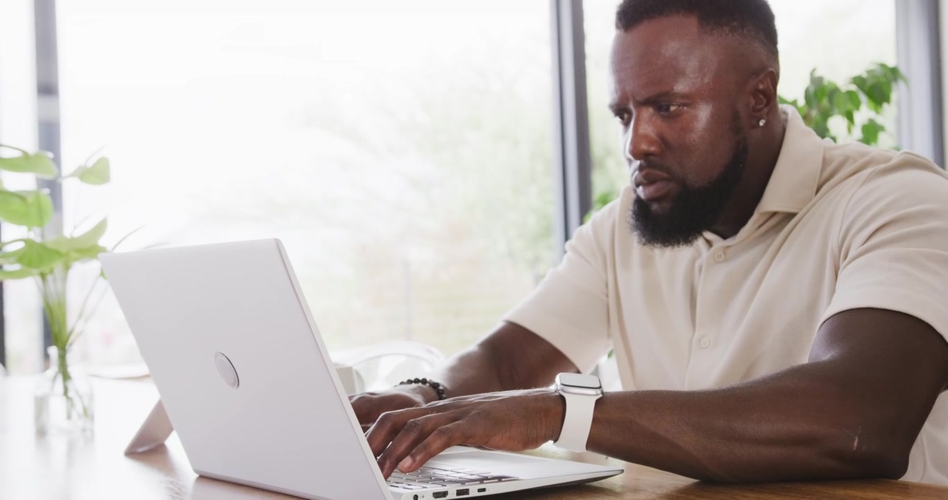 Man typing on white laptop at wooden table by window with plants, wearing polo and smartwatch