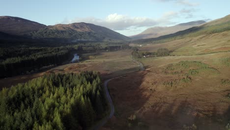 aerial drone footage of a winding single track road stretching out towards mountains, surrounded by a forest of conifer trees, moorland and a river