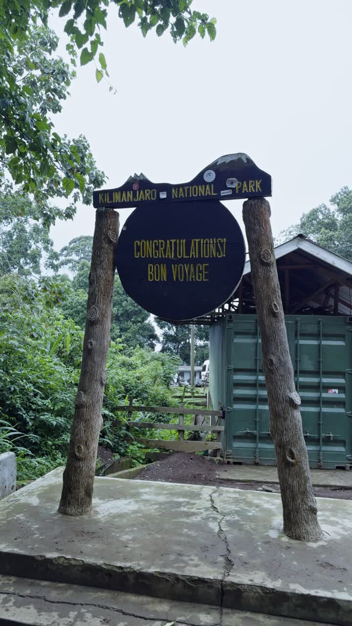 Vertical shot captures the round Congratulations Bon Voyage sign at Mweka Gate exit in Mount Kilimanjaro National Park, set against lush trees and a trail