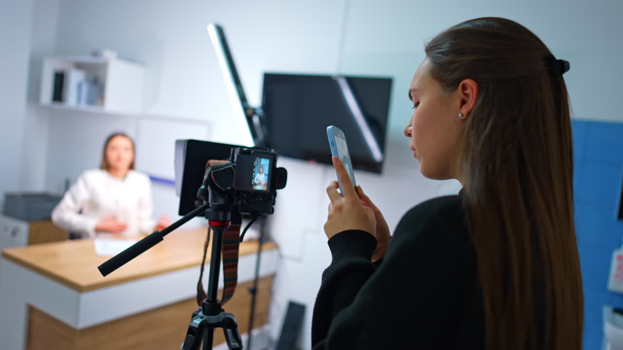 Brunette girl takes video on her phone of the display of professional camera. Female blogger speaks to camera at blurred backdrop.
