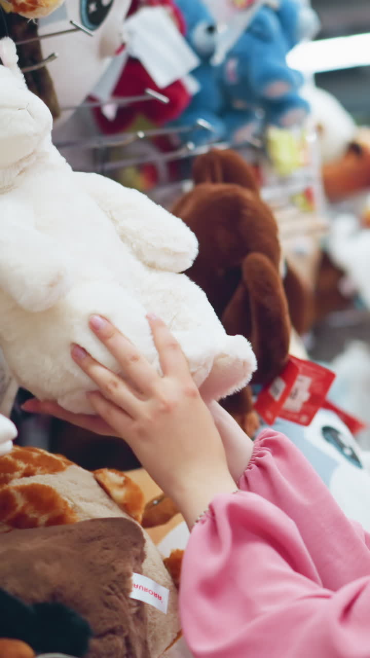 분홍색 드레스를 입은 여자가 장난감 가게에서 색 플러시 장난감을 검사하고, 집중하고 검사합니다. 그녀를 둘러싸고 있는 다른 다채로운 은 장난감은 밝게 밝은 가게의 선반에 끔하게 배열되어 있습니다.