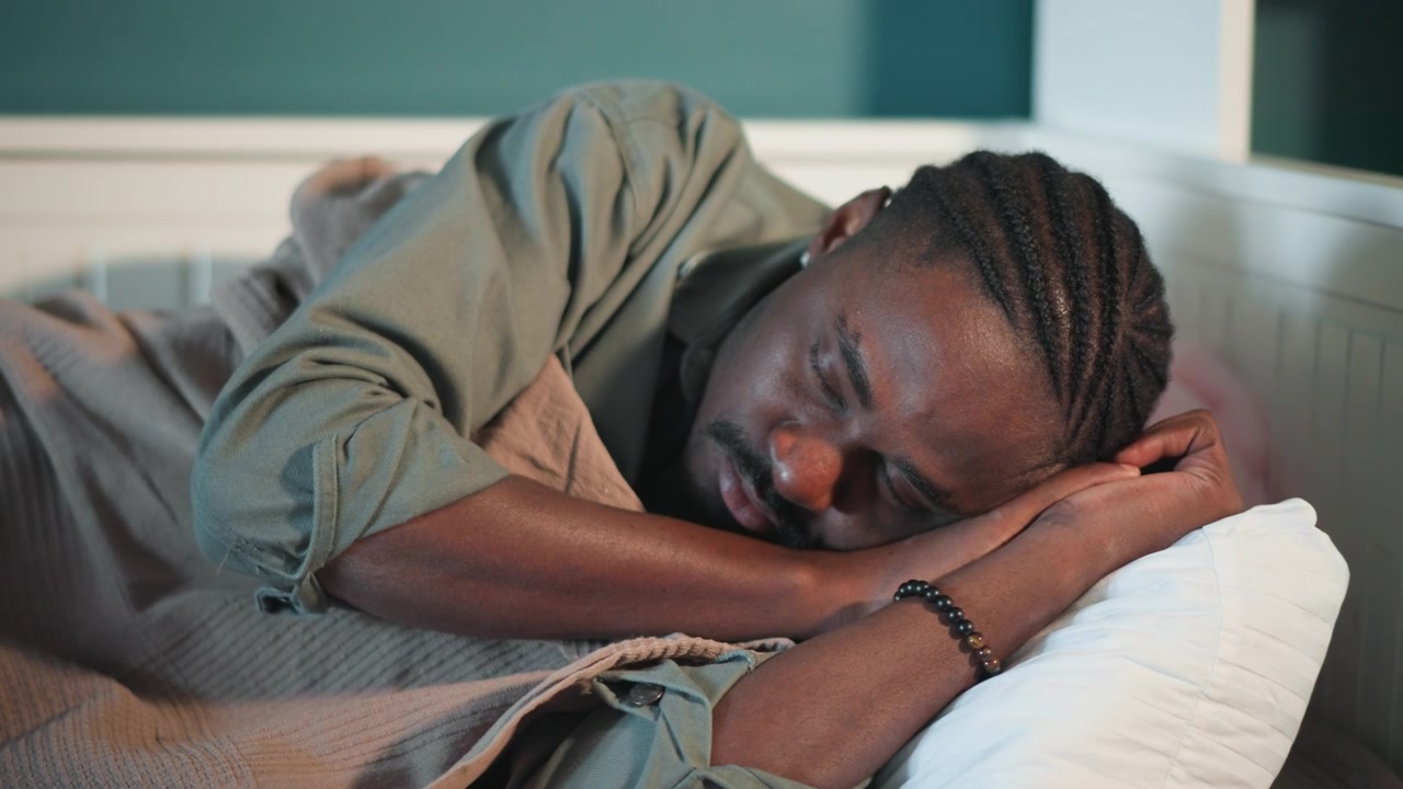Tense man waits anxiously. Man with braided hair experiences feverish discomfort in quiet room. Individual with braided hair exhibits signs of fever while waiting in dimly lit bedroom
