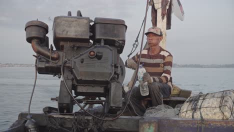 an exhausted old fisherman sips water while steering his aged boat, powered by an old engine, homeward-bound