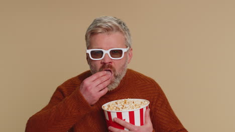 un hombre con un suéter marrón y barba lleva gafas 3d y sostiene un cubo de palomitas de maíz, sonriendo emocionado.