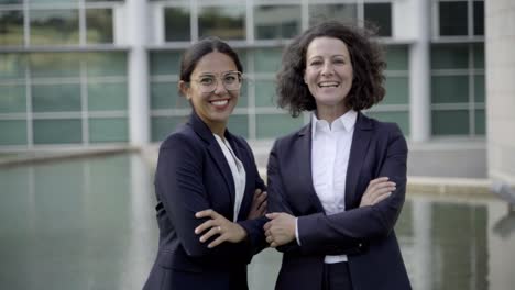 mujeres sonrientes con los brazos cruzados de pie cerca del edificio de oficinas
