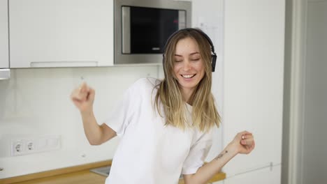 Una-Joven-Feliz-Bailando-En-La-Cocina-Vistiendo-Pijamas-Blancos-Y-Escuchando-Música-Con-Auriculares.