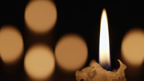 wax burning candle flickers in dark on a black background with blurred candlelight bokeh effect. background or illustration of remembrance, mourning or celebration. blaze candles in church close up