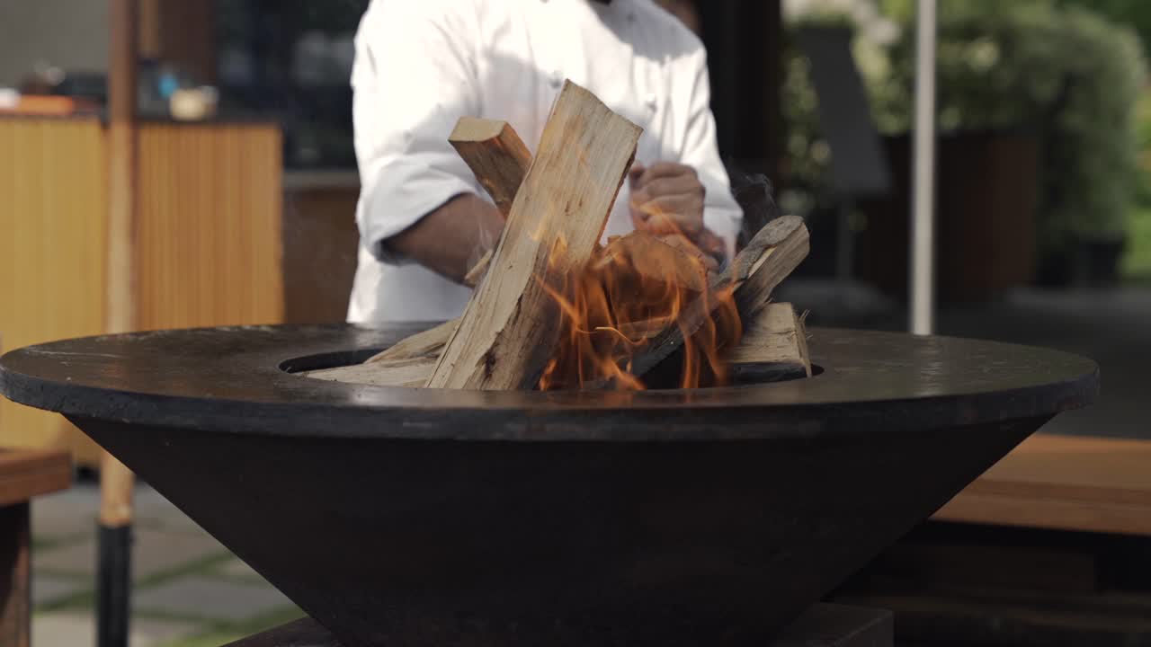 A chef in a white uniform ignites a wood fire brazier brasero in an outdoor fire pit, preparing for open-flame cooking. The flames flicker around stacked logs, creating a rustic ambiance.