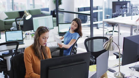 dos mujeres de negocios caucásicas sentadas en escritorios usando computadoras girándose para hablar entre sí