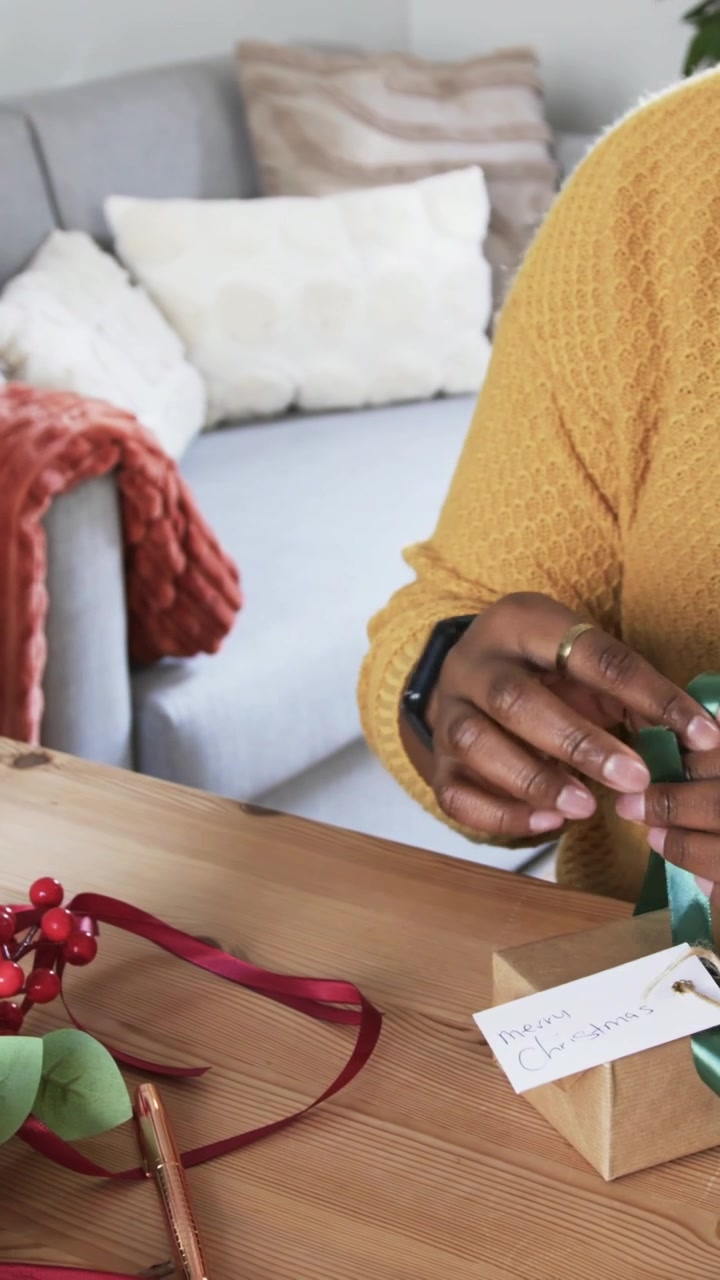 Vertical video: Picking tag, woman in yellow sweater tying kraft gift box at home with green ribbon