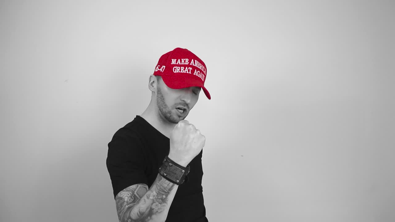 Grayscale footage of a man with tattoos, short hair and a beard who is wearing a red colored MAGA Make America Great Again hat celebrating a victory or something by screaming and lifting up his arms.