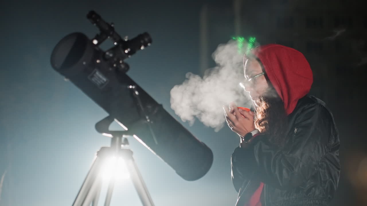Osservazione notturna con telescopio, alito freddo visibile, astronomo amatoriale con felpa rossa scalda le mani con il respiro accanto a un grande telescopio riflettore su treppiede sotto la luce urbana con una leggera retroilluminazione e lampada