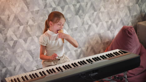 kid with long brown hair in white blouse sitting by keyboard, holding and enjoying large ice cream cone with joyful expression, indoor setting with soft patterned wall and beanbag in background